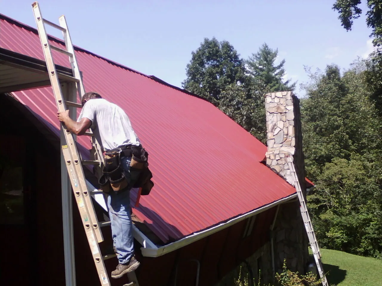 Roofer on a ladder beside a red metal roof and stone chimney — Soffit & Fascia Repair by Roofing Brothers in Walhalla